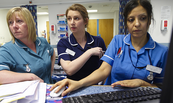Three nurses stand together at ward station. They appear busy