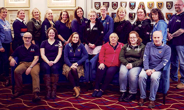 Members of the Veterans Clinical Services team, who won the Community and General Practice award at the RCN Nursing Awards 2023, sitting and standing in a semi-circle a room with service shields and pictures on the wall behind them