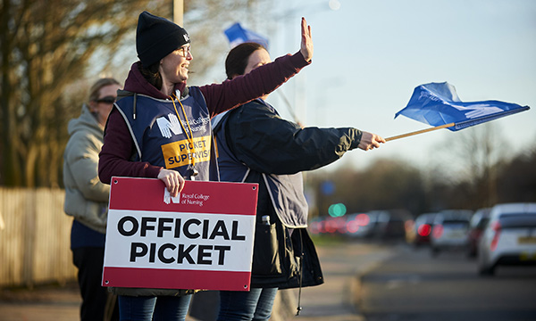 NHS striker holding RCN picket sign waves at unseen person. Road in front of her is traffic-lined